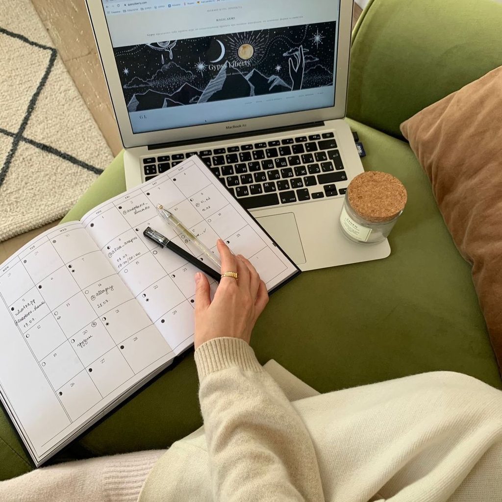 A young adult writing realistic goals in a notebook calendar with a laptop